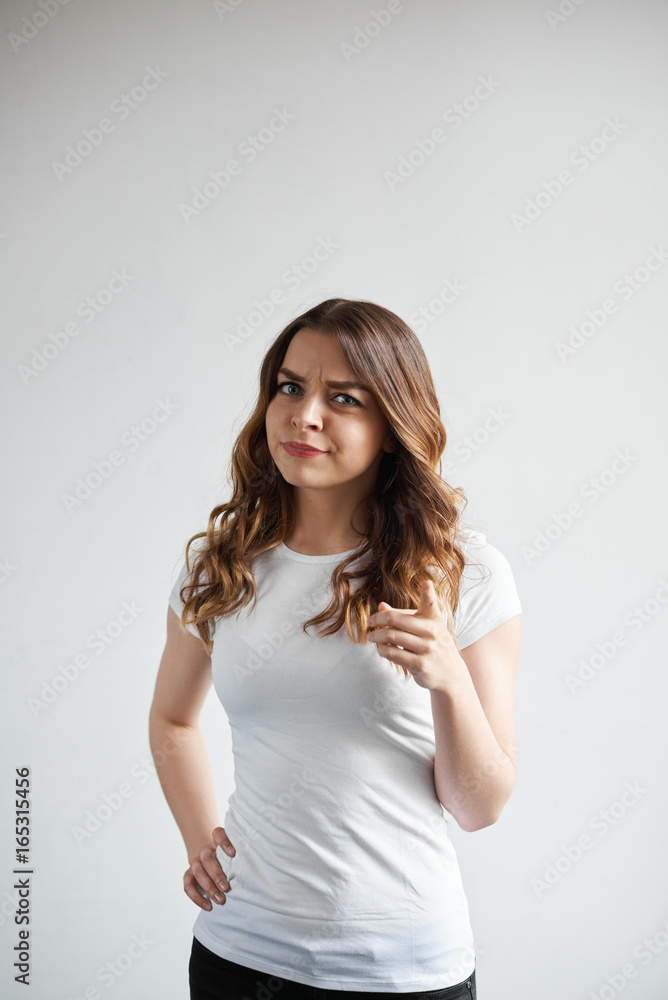 Attractive young woman with long wavy hair frowning and pointing finger at camera, having displeased grumpy expression, warning or reproaching someone. Human emotions and body language concept Attractive young woman with long wavy hair frowning and pointing finger at camera, having displeased grumpy expression, warning or reproaching someone. Human emotions and body language concept