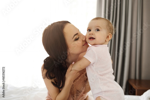 Portrait of young happy tender mom and her baby daughter smiling sitting on bed at home.