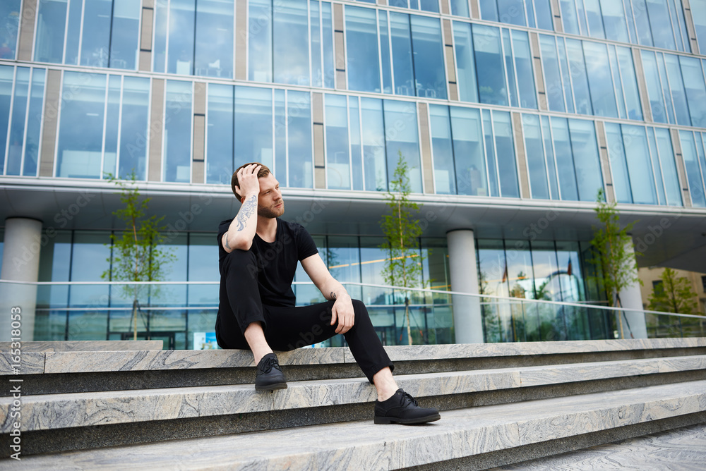 People and modern urban lifestyle concept. Handsome tattooed young guy wearing trendy black clothing sitting on concrete stairs against office building background and waiting for his girlfriend