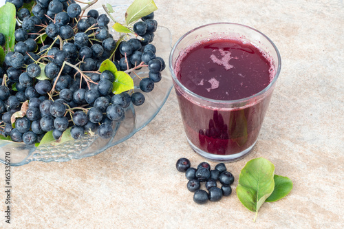 Fresh juice of black chokeberry (Aronia melanocarpa) in glass and berry in pot on brown ceramic background