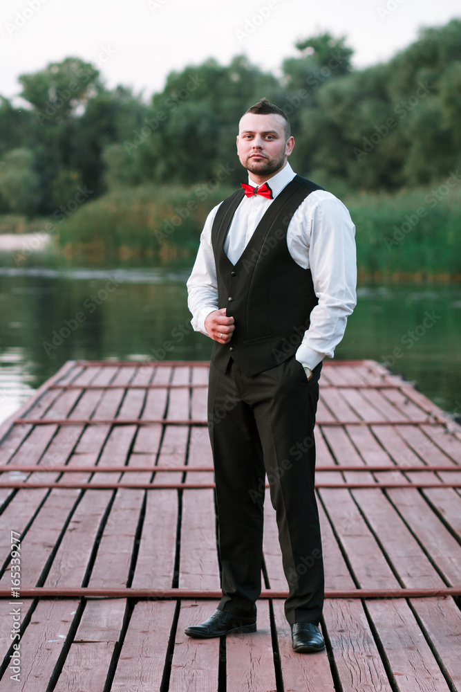 Young stylish man in a waistcoat, vertical portrait of the groom, portrait on a background of nature, the river and the pier, look at the camera Young stylish man in a waistcoat, vertical portrait of the groom, portrait on a background of nature, the river and the pier, look at the camera