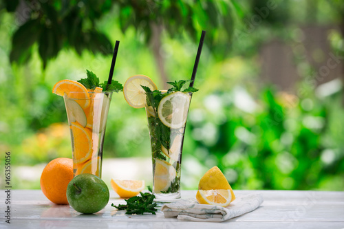 Detox water with fruits - orange and green apple and lemon and basil leaves and mint leaves on white table outdoors in summer garden. Homemade mojito or lemoade with ice cubes. photo