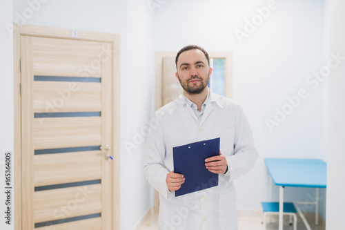 Cheerful young doctor holding a clipboard and gesturing with his hand at hospital ward
