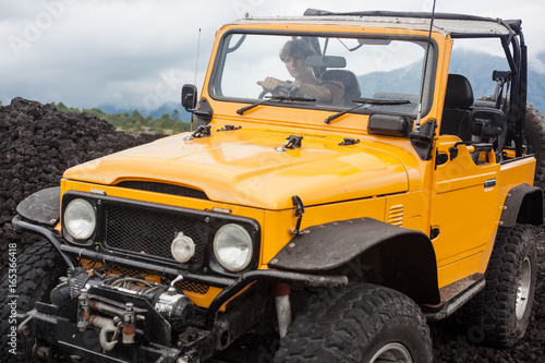 A curly-haired man is looking away in the offroad yelow vehicle at the top of a valley with volcanic rock and mountains in Bali, Indonesia