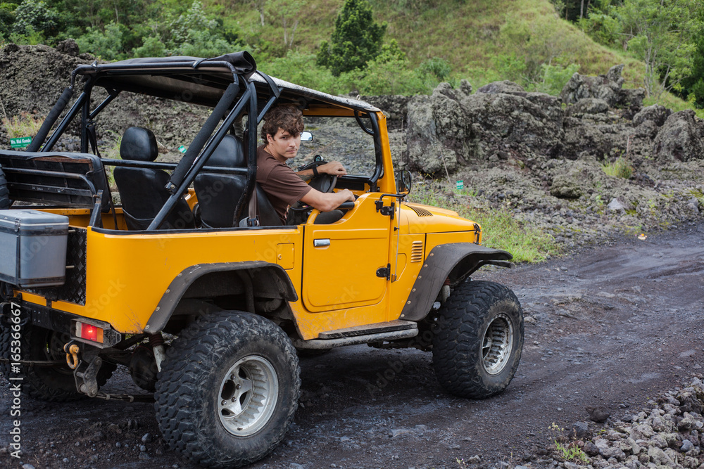 Side view of a driver looking away and sitting in yellow offroad vehicle parked on a road with dark ground among green trees.