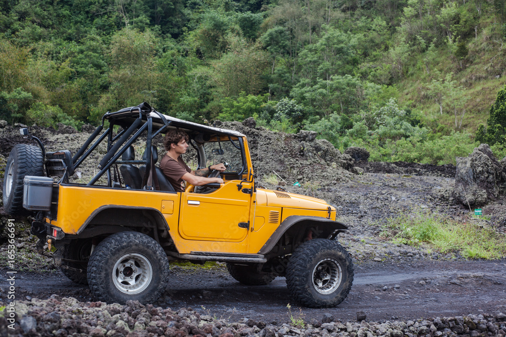 Side view of a driver watching forward and sitting in yellow offroad vehicle parked on a road with dark ground in front of the forest. Side view of a driver watching forward and sitting in yellow offroad vehicle parked on a road with dark ground in front of the forest.