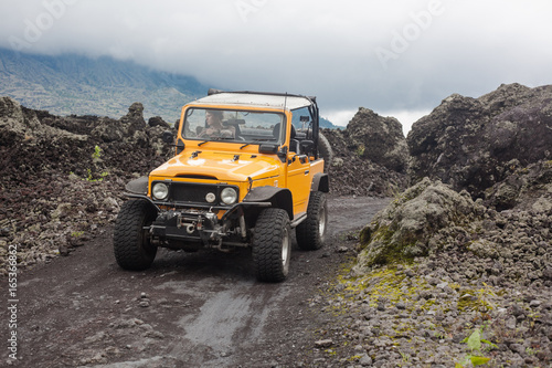 A curly-haired man is looking away driving an offroad yelow vehicle at the top of a valley with dark ground road