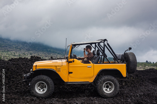 A curly-haired man drinking water is sitting in the offroad yelow vehicle parked at the top of a valley with volcanic rock and mountains in Bali, Indonesia