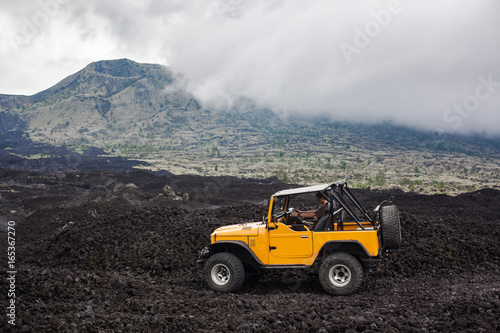 A curly-haired man is sitting in the offroad yelow vehicle parked at the top of a valley with volcanic rock and mountains in Bali, Indonesia