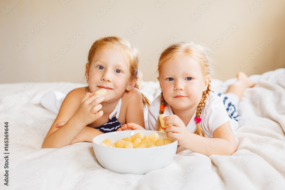Portrait of two white Caucasian children girls eating corn puffs. Sisters eating snack fast food in bed at home indoors. Healthy meal childhood concept.