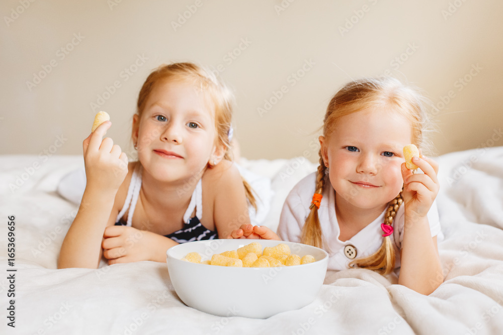 Portrait of two white Caucasian children girls eating corn puffs. Sisters eating snack fast food in bed at home indoors. Healthy meal childhood concept. Portrait of two white Caucasian children girls eating corn puffs. Sisters eating snack fast food in bed at home indoors. Healthy meal childhood concept.