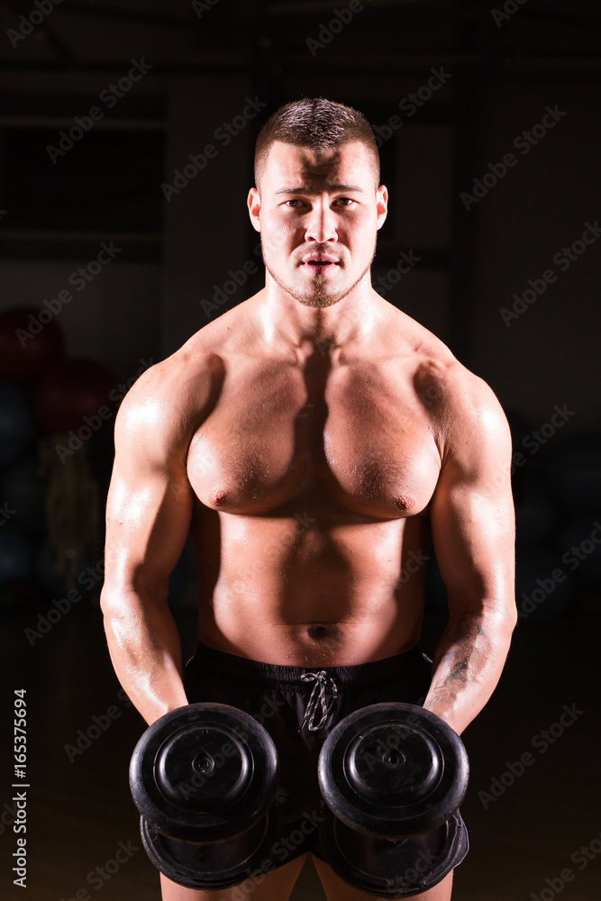 Handsome power athletic guy bodybuilder doing exercises with dumbbell. Fitness muscular body on dark background