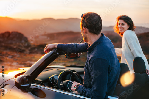 Quadro su tela A young couple have fun in a convertible at sunset, around them beautiful nature