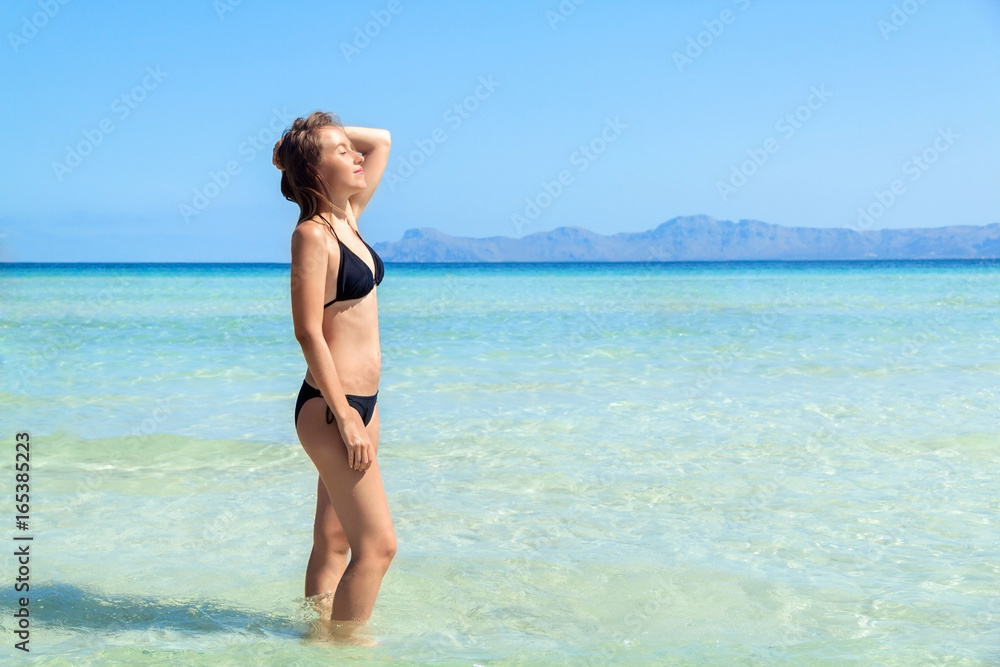 Young woman in bikini standing with her hand over head enjoying hot summer day on the beautiful beach of Alcudia, Mallorca, Spain Young woman in bikini standing with her hand over head enjoying hot summer day on the beautiful beach of Alcudia, Mallorca, Spain