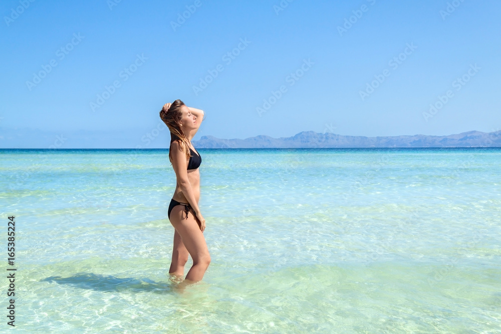Young woman in bikini standing with her hand over head enjoying hot summer day on the beautiful beach of Alcudia, Mallorca, Spain