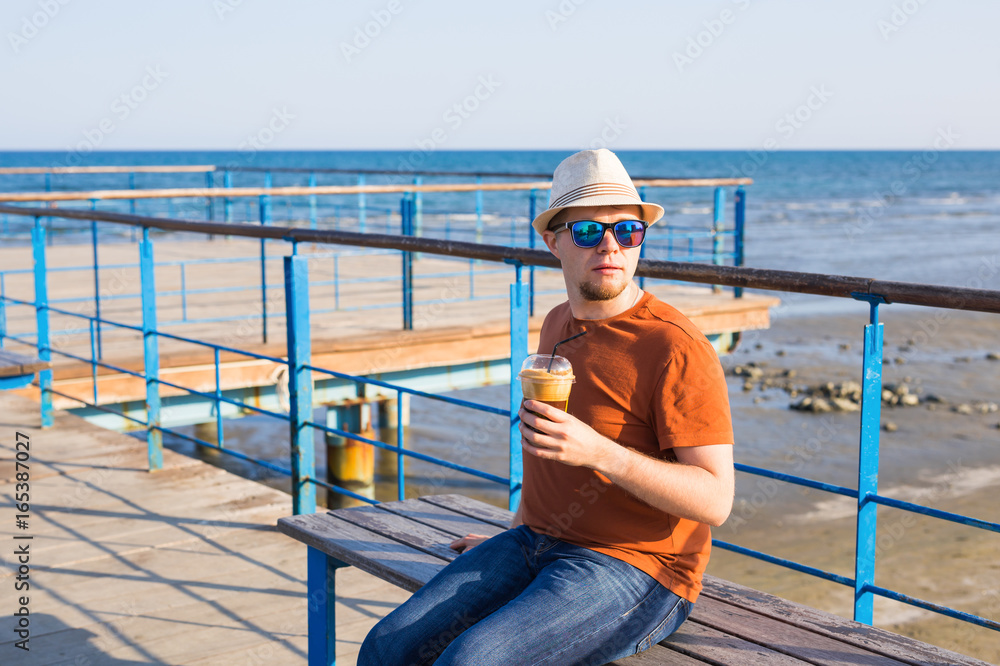 Portrait of cheerful attractive bearded hipster young man drinking iced coffee or frappe on the background of the sea