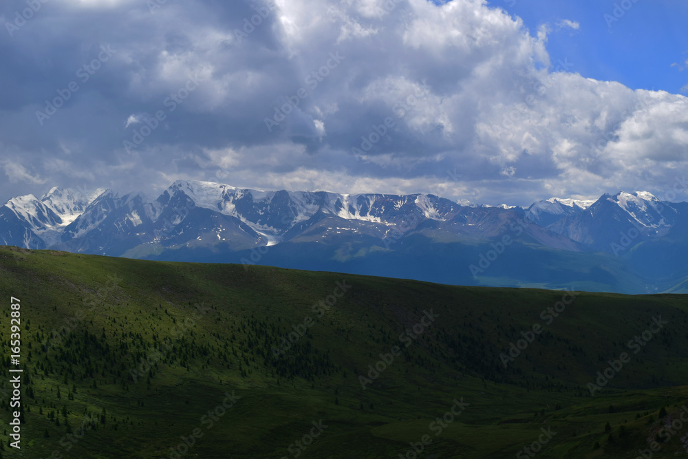 View of white peaks of North-Chuyski ridge and hills with green fields in Altai mountains in cloudy weather. Aktash, Altay Republic, Russia.