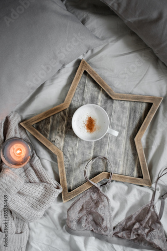 A cup of coffee and a candle in a Scandinavian wooden tray in a cozy bed with pillows.