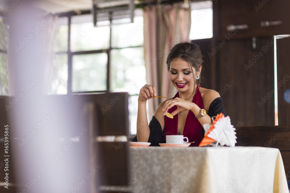 Young business woman grinks coffee at a cafe. Smiling and flirts. Looks at camera