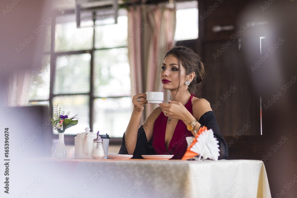 Young business woman grinks coffee at a cafe. Smiling and flirts. Looks at camera Young business woman grinks coffee at a cafe. Smiling and flirts. Looks at camera