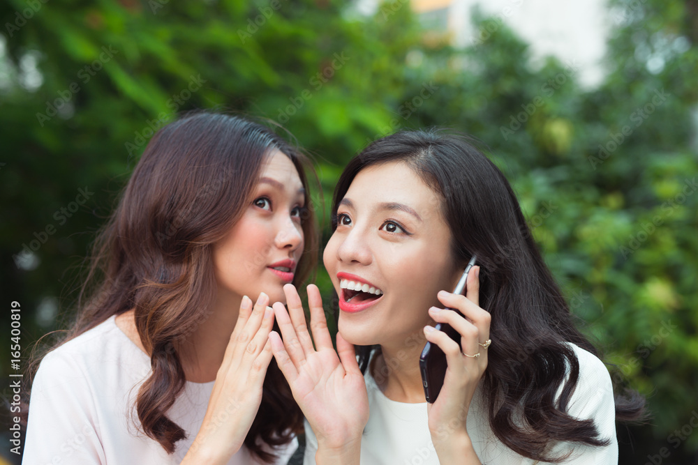Young woman whispering into cheerful friend's ear while on call Young woman whispering into cheerful friend's ear while on call