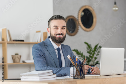portrait of smiling businessman looking at camera at workplace with laptop