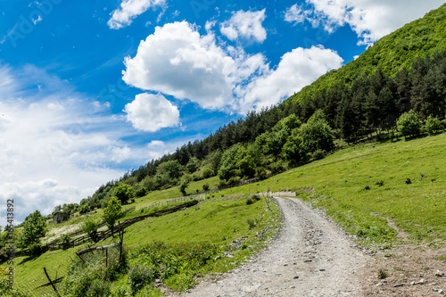 Slope of the Alpine meadow with a picturesque gravel road against a blue sky with white clouds
