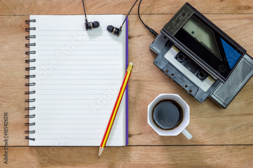 Bank notebook with pencil laying on the brown table. Vintage old tape player with earphones and black coffee put on the table as well.