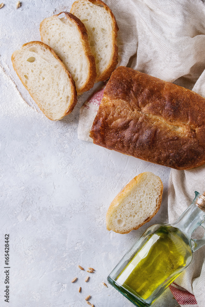 Homemade white wheat bread whole and slice served with bottle of olive oil and wheat grain seeds on white linen towel over gray texture background. Top view with space. Homemade white wheat bread whole and slice served with bottle of olive oil and wheat grain seeds on white linen towel over gray texture background. Top view with space.
