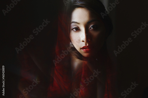 Bride holds her chin on the shoulder standing behind a red curtain © IVASHstudio
