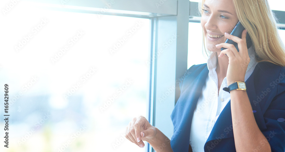 Businesswoman standing against office window talking on mobile phone Businesswoman standing against office window talking on mobile phone