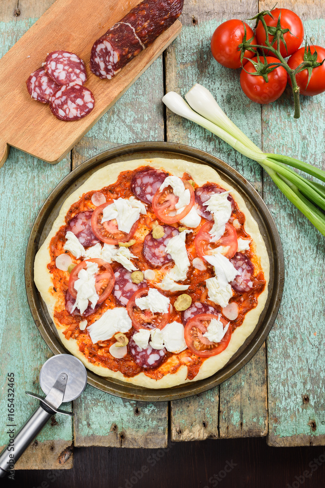 Raw rustic pizza with smoked salami, tomatoes, green olives and cheese served with pizza knife and ingredients: leek, cherry tomatoes and salami