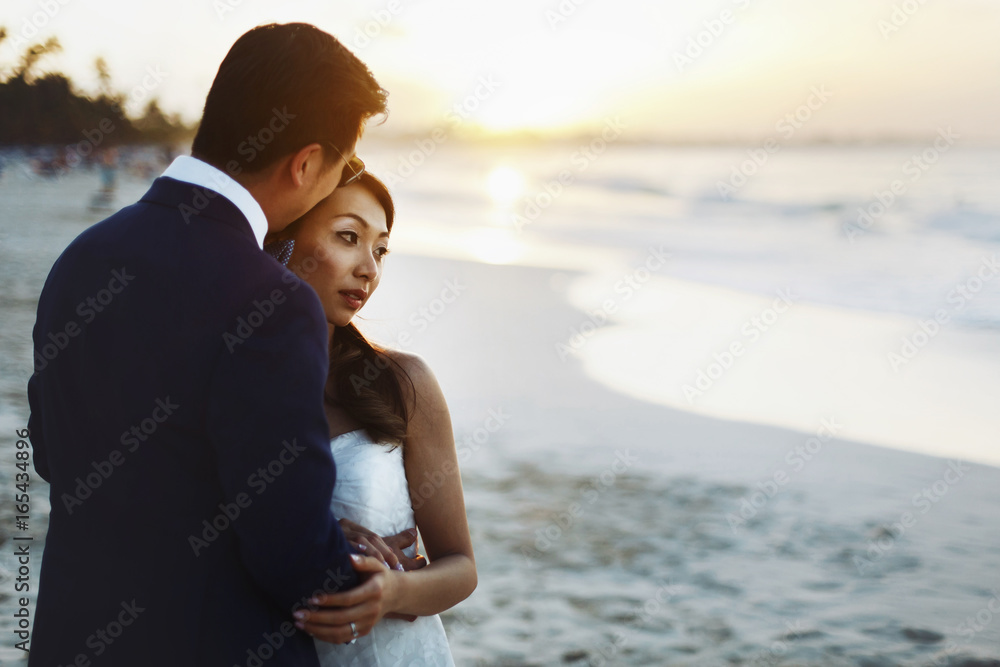 Bride and groom hug each other tender standing in the rays of golden sun on the beach Bride and groom hug each other tender standing in the rays of golden sun on the beach