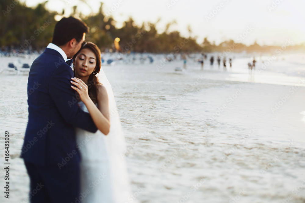 Bride and groom hug each other tender standing in the rays of golden sun on the beach Bride and groom hug each other tender standing in the rays of golden sun on the beach