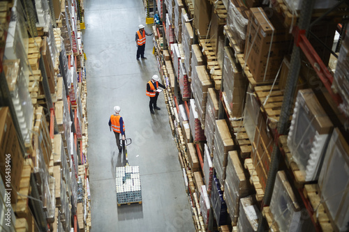 Above view of warehouse workers moving goods and counting stock in aisle between rows of tall shelves full of packed boxes