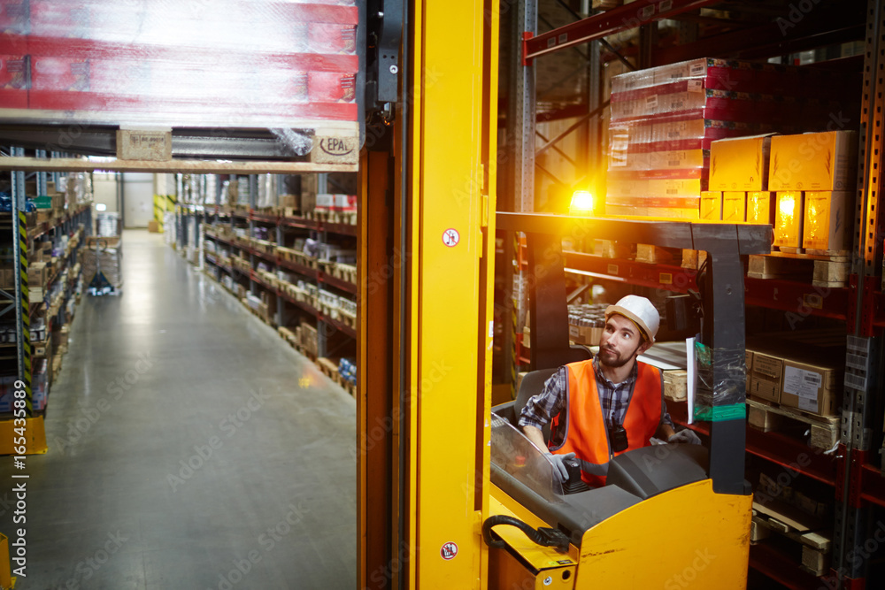 High angle portrait of warehouse worker using reach fork truck to load pallet with boxes on tall rack