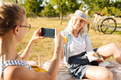 Pleasant teenager girl taking photos of her grandmother on a picnic