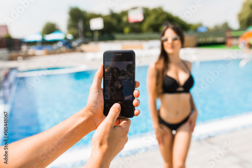 Hand with phone take photo of beauty sexy woman in swimwear near swimming pool. Time for summer photo.