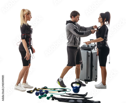 Trainer helps fitness women to set full electrical muscular stimulation suits. Various sport gear like kettlebells  dumbbells  belts and expanders around them.