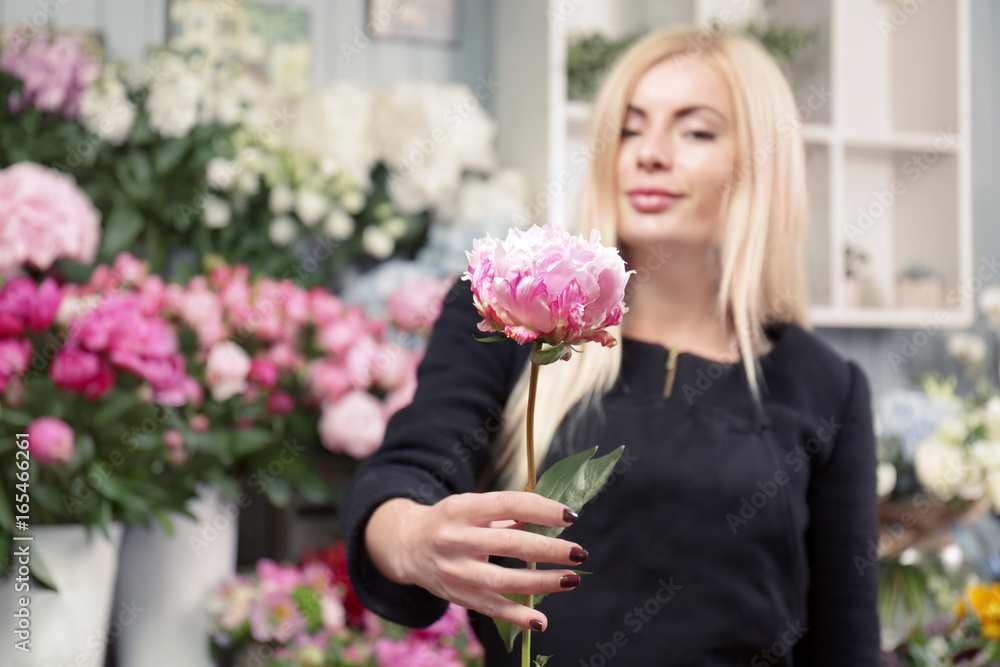 Fototapeta premium Blonde florist presenting fresh pink peony flower in a retail flower shop with blurred floral background, the flower is in sharp focus, its delicate petals and vibrant color