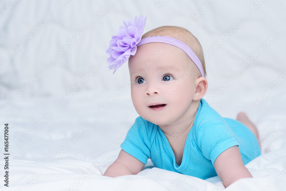Cute baby girl in blue shirt, with bow flower on head, lying on stomach on soft blanket and looking with surprise, indoors Cute baby girl in blue shirt, with bow flower on head, lying on stomach on soft blanket and looking with surprise, indoors