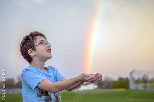 Young boy looking up in awe at bright rainbow after spring rain, trying to catch the colors in his hands
