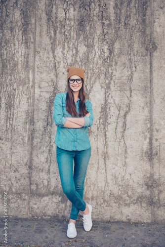 Full length of excited girl, standing on the concrete wall`s background outdoors, smiling, with crossed legs and hands, in casual jeans outfit and brown hat, glasses