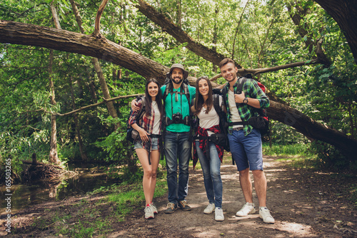 Close up of four cheerful friends in the summer nice wood. They are hikers, walking and picking place for camping, embracing, posing for photo with equipment