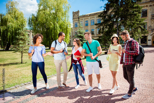 Free time of a students, bachelor`s campus life rhythm. Six friendly students are walking after they passed test outside the college building and discuss the project, smiling, enjoying, carefree