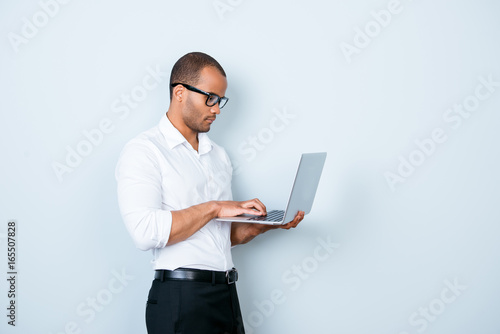 Confident pensive young americano market broker is looking at his laptop, standing on the pure background, being pensive and severe