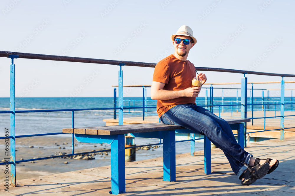 Portrait of cheerful attractive bearded hipster young man drinking iced coffee or frappe on the background of the sea Portrait of cheerful attractive bearded hipster young man drinking iced coffee or frappe on the background of the sea