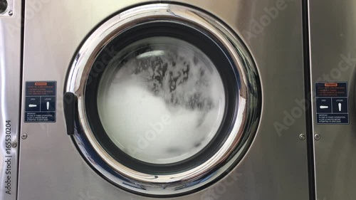 4K HD video of laundry agitating in a large industrial sized washing machine, stopping then re-starting. Bubbles forming viewable through door window.