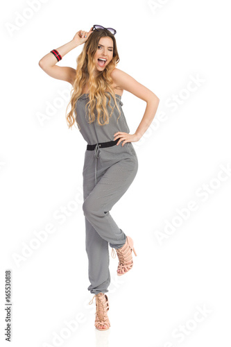 Happy Young Woman In Jumpsuit Is Standing On One Leg And Winking
