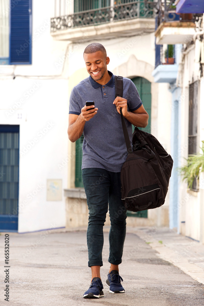 Fototapeta premium smiling young black guy traveling with bag and using mobile phone on the street